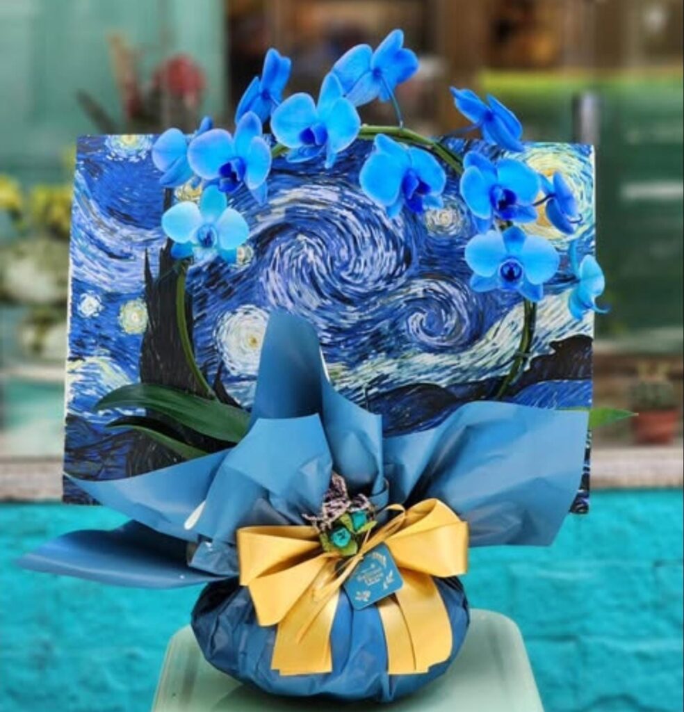 Orquídea azul em arranjo artístico inspirado em “Noite Estrelada”, com embalagem azul, laço amarelo e painel decorativo ao fundo.