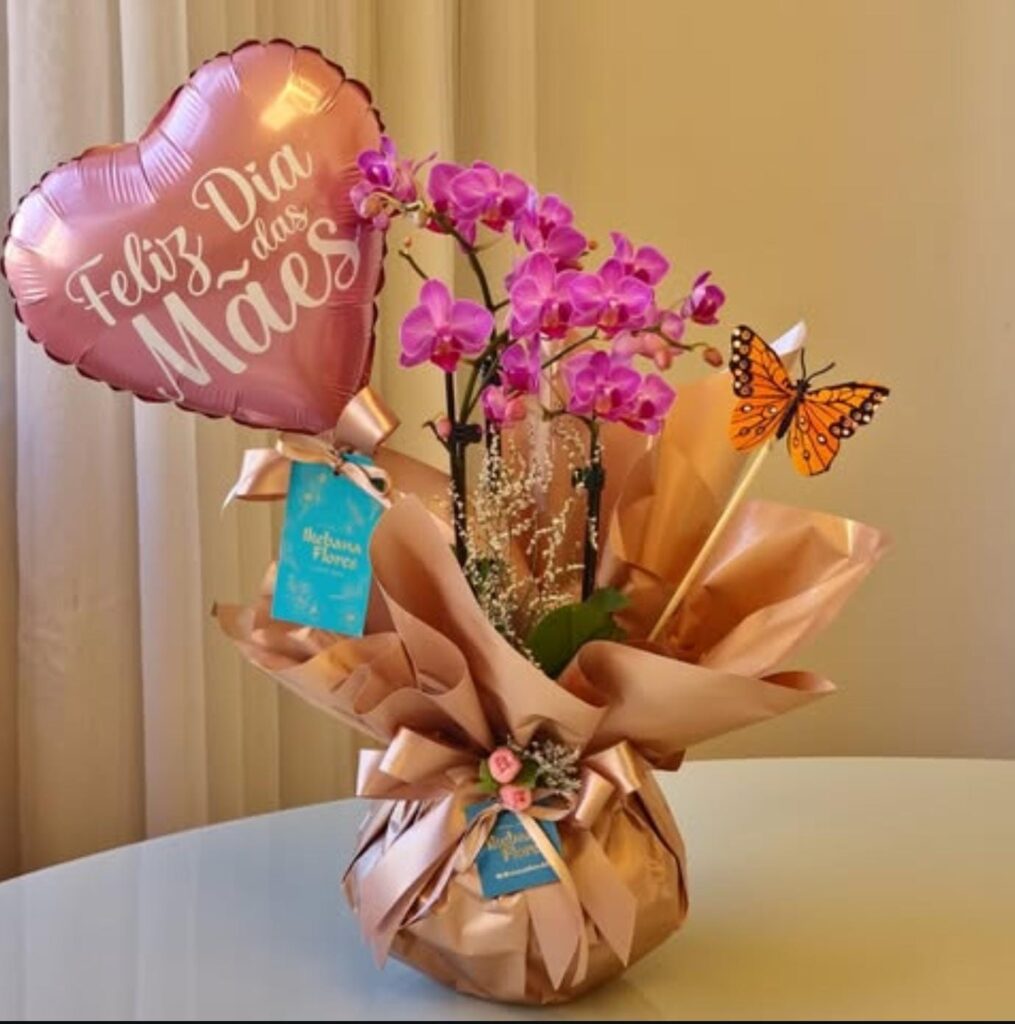 Orquídea rosa em embalagem sofisticada rosé, acompanhada de balão de coração “Feliz Dia das Mães” e borboleta decorativa, sobre mesa em ambiente interno.